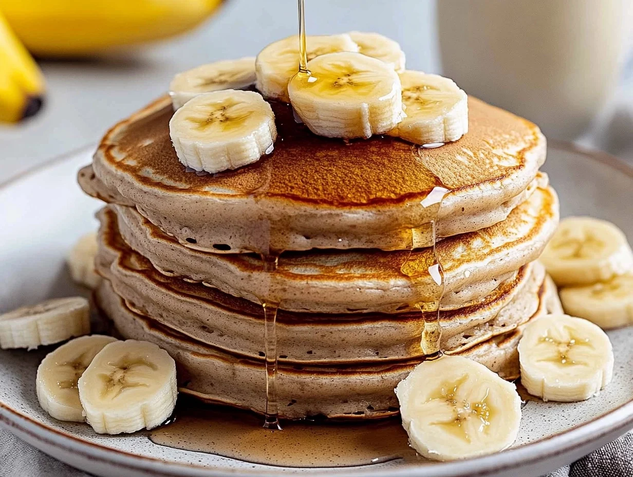 Stack of fluffy banana pancakes topped with fresh banana slices and drizzled with maple syrup on a white plate.